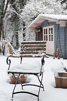 A snow covered seat, with view to blue wooden summerhouse. 
