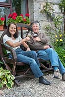 Couple enjoying a glass of wine on a rustic wooden carriage wheel seat. 
The front of a stone house with climbers and windowboxes of flowers