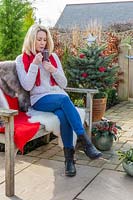 Woman relaxing on bench with sheepskins, a decorated Christmas tree in background having a hot drink. 
