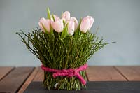 Pale tulips displayed in a vase decorated with Vaccinium - Bilberry branches tied around a jar. 