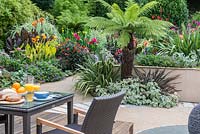 Intimate dining area on a deck overlooking a courtyard edged in beds of Australian tree ferns, cannas, phormiums, dahlias, hardy geraniums and silver Plectranthus.
