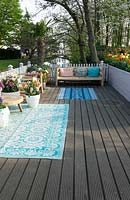Wooden terrace furnished with wooden bench, cushions, rugs and pots of spring flowering perennials. 