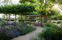 View up flowering border and pathway to pleached trees, wooden pergola and dining area. 