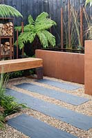 A chunky oak bench, Corten water trough and log storage in a contemporary, urban style garden in 'The Penumbra' garden, RHS Tatton Park Flower Show 2018