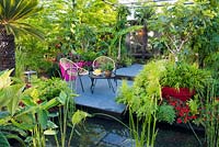 Seating area in the garden with tender planting in pots by the pond with aquatic plants Equisetum hyemale, Cyperus and Thalia. B and Q Bursting Busy Lizzie Garden at RHS Hampton Court Palace Garden Festival Show 2018