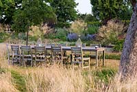 Table and chairs set in shade of apple trees, surrounded in uncut grass. Beyond borders studded with large clumps of Stipa gigantea.