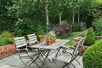 A small paved courtyard with dining table and chairs, is hidden away behind a tall hornbeam hedge and silver birches, Betula utilis var. jacquemontii.