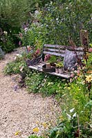 Gardening tools and cushion on Rustic garden bench nestled amongst perennial planting along winding gravel path. RHS Hampton Court Festival 2019. 