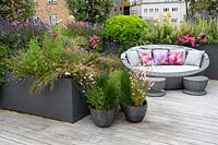 Roof terrace with wood decking and garden sofa with built in shade and decorative cushions, with perennials and annuals forming a natural screen for privacy. Planting includes Pittosporum tobira Nanum, Gaura lindheimeri Whirling Butterflies, Juncus maritimus, Verbena bonariensis, Pink Bedding Geraniums, Olea europaea, Geum Totally Tangerine.
