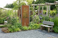 Small garden with gravel mulch, wooden pergola and bench with words meaning 'nice and sustainable'. A steel rusted screen with tree design. Mixed informal planting including Carex morrowii and Agastache. 