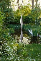 The Savills and David Harber Garden - View of the pool with the sculpture representing a leaf and surrounded with a woodland clearing, the planting includes Anthriscus sylvestris, Tellima grandiflora, Iris pseudacorus, and Ranunculus acris - Sponsor: Savills and David Harber.