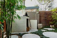 Pizza oven in back corner of garden, next to a painted brick fence, a timber slat screen with an outdoor shower, a stone carved Indonesia pot, a family group of sculptures, a large glazed water bowl, semi rustic stone pots, concrete circular cast in situ stepping stones in amongst a thick planting of mini Mondo grass.