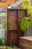 Freestanding hardwood timber slat screen with an inbuilt cold water tap next to a timber bench seat infront of a painted brick fence, featuring a Rhipsalis, succulent in a hanging basket.