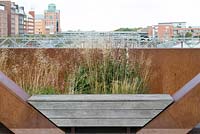 Wooden bench between corten steel wall with view of the railway and city buildings.