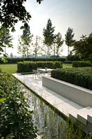 Long rill alongside paved patio with dining area in modern garden. 