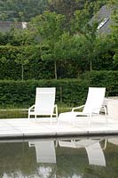 Recliners on paved patio surrounded by pool in modern garden. 