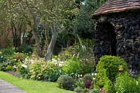 View of mixed borders with trees underplanted with perennials and bulbs, stone outbuilding nearby
