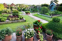View of kitchen garden from terrace edged with container plants. Kitchen garden divided into quarters by paved paths, each quarter has lawn with large raised beds edged with railway sleepers, blue arbour along boundary fence
