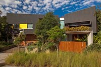 A wide view of a contemporary style house with small trees, a kerbside planting of grasses and a suspended garden planted with bromeliads.