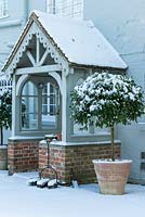 Pair of terracotta pots with Laurus nobilis - standard Bay trees by Wooden and brick porch with boot scraper and brushes, snow in February. The Old Rectory, Suffolk, UK