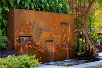 Freestanding rusty corten steel fountain, featuring internally lit laser cut patterns representing mountains and clouds, with three rills behind pale grey stepping stones.