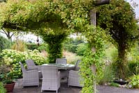 Rose covered arbour over a dining area in a rural garden with bubble millstone water feature in the foreground and grasses and hydrangea behind.