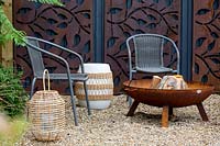 With gravel patio, and chairs around a fire pit. In background decorative rusty metal panels on wooden fence.