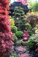 Red buddha statue set amongst autumnal colours of mixed acers, conifers, cloud tree and azaleas with black Japanese tea house behind, at Four Seasons garden, Walsall, West Midlands.