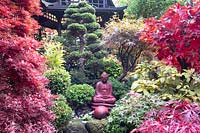 Red buddha statue set amongst autumnal colours of mixed acers, conifers, cloud tree and azaleas with black Japanese tea house behind, at Four Seasons garden, Walsall, West Midlands.
