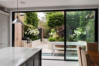 View from kitchen diner through glass sliding doors onto porcelain paved garden with outdoor dining area and raised beds.