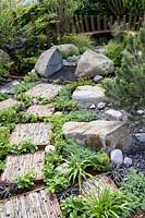 Natural rock and stone, with green foliage groundcover planting. Wooden seating and  stepping stones with slate pieces in corten steel frames with Deschampsia cespitosa, Achillea 'Moonshine', Ophiopogon planiscapus 'Nigrescens' - Through Your Eyes Garden - RHS Hampton Court Palace Garden Festival 2019. Sponsors: Kebony, CED Stone, R and G Metal Products, William's Art and Design, Practicality Brown.