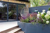 London contemporary garden with view towards house - bespoke barbecue on patio with grey raised border. Planting includes Heuchera berry smoothie, Salvia caradonna, Hydrangea anabelle, Geranium johnsons blue.