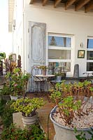Corner of a verandah with rusty metal outdoor table and chairs and potted, mismatched a retro metal bucket with a succulent and a metal tub with a Pelagonium in it in front of a shabby chic panelled door.