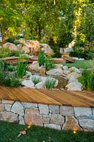 A natural looking stream and waterfall constructed from large boulders and small rocks, next to a small timber deck, interplanted with a variety of flowering and strappy leaved plants, featuring a low arched timber bridge and a small lawn area with circular seagrass cushions.