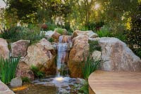A waterfall constructed from large boulders and small rocks, planted out with grasses, flowering plants and groundcovers and featuring a curved hardwood timber boardwalk