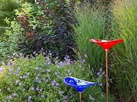 Two glass bird baths in front of mixed bed with Panicum virgatum 'Shenandoah' and Geranium 'Rozanne', Continus coggygria in background