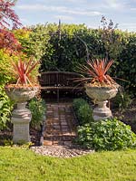 Path of recycled bricks leads to wooden bench under metal arbour, two urns o plinths either side of the path planted with Cordyline australis 'Torbay Dazzler' 
