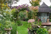 Sixth-of-an-acre, irregular plot has an arch clad in Rosa 'Veilchenblau' framing view of herbaceous borders. A thatched  summerhouse and deck overlook a bog garden and pond. To left, Ammi majus stands beneath magnolia.