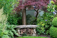 A small stone bench with logs stored beneath, is partly shaded by a Japanese acer.