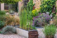 A salvaged galvanised water trough is converted into a water feature, and planted with irises and water lilies. Clumps of blue fescue and miscanthus grow in the gravel.