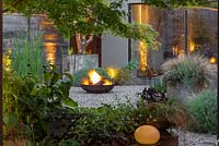 Seen through a window of foliage, a flaming fire pit reflected in the house windows.