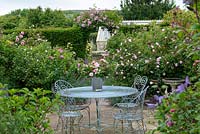 Table and chairs on paved area surrounded by roses such as Rosa 'Jacques Cartier' and 'Fantin Latour'.  Rosa 'Laure Davoust' on arch.