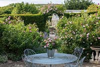 Table and chairs on small paved area with Rosa 'Jacques Cartier' and  'Fantin Latour'. Rosa 'Laure Davoust' over arch.