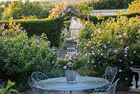 Table and chairs in classic rose garden with Rosa 'Jacques Cartier' and  'Fantin Latour'. Rosa 'Laure Davoust' over arch.