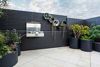 View from in doors to outside rooftop garden featuring a raised garden bed groups of grey cylindrical pots and a wall mounted barbecue.