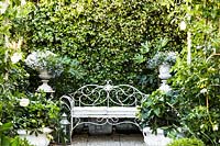 Ornate metal bench seat in courtyard garden with classic urn containers on plinths 