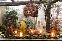 Outdoor table decoration made with Corylus avellana 'Contorta',  and branches of Abies nordmanniana, Picea pugnes 'Hopsii', Pittosporum tobira berries, pine cones, candles and mandarins