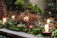 Outdoor table decoration made with 'twisted' hazel branches of Corylus avellana 'Contorta', branches of Abies nordmanniana, Picea pugnes 'Hopsii', Pittosporum tobira berries, rosehips, candles pine cones and mandarins