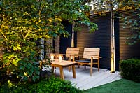 Illuminated seating area with lanterns surrounded by Cornus kousaÂ var.Â chinensis and pleached Quercus ilex by black wooden fence