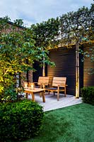 Illuminated seating area with lanterns surrounded by Cornus kousaÂ var.Â chinensis and pleached Quercus ilex by black wooden fence.